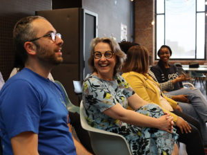 un homme assis avec un tee-shirt bleu et des lunettes et une femme assise à côté entrain de sourire